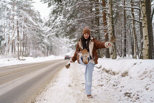 Joyful young Asian woman travel blogger or photographer in stylish sheepskin coat kicking snow towards camera and laughing joyfully while walking in winter nature, hitchhiking along snowy forest road