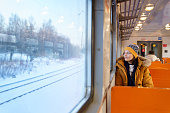 Pensive young European woman wear hat traveling by local train in winter time, thinking, looking through the window at snowy landscape.