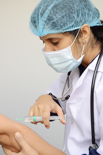 Vertical photo of a woman nurse or doctor or health care worker, physician injecting vaccine with syringe in her hand to administer a dose over gray background with copy space for text.
