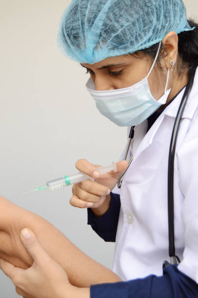 a woman or lady or female physician doctor or nurse in surgical mask, cap, and white lab coat and a stethoscope around her neck holding a syringe needle injection in her hand to administering shot dose to a patient in the arm , over grey background - syringe medical injection surgical needle surgical mask ストックフォトと画像