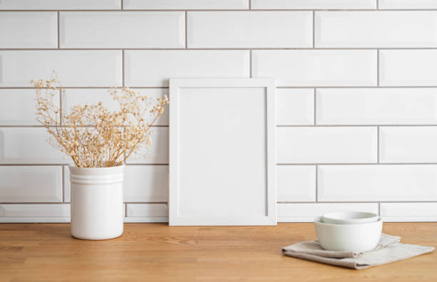 istock A  frame with a blank canvas against a white tile wall and on a wooden tabletop with vase and cup. 1966729497