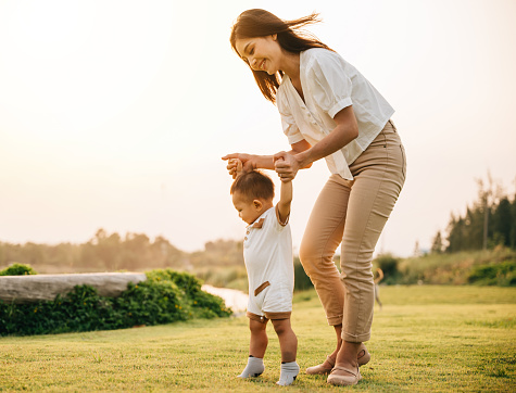 A beautiful Asian family enjoying a sunny day in the park as they teach their baby to take his first steps. Cute toddler little child family concept