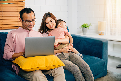 In their home office a husband works on his laptop while his wife carries their sleeping daughter epitomizing delicate balance between family work and responsibility. Love and connection are evident.