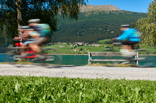 3 cyclists in motion. Adults doing sports with bicycle on sunny summer afternoon, at the lake in the mountains. Italy, Vinschgau, Lake Resia (Reschensee).