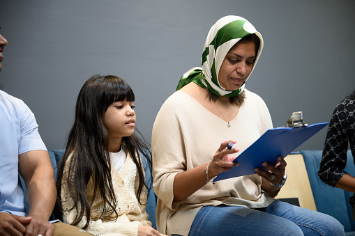 Mother wearing headscarf fills in medical questionnaire sitting with family in waiting room