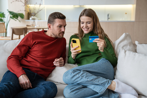 Online shopping. Overjoyed wife with credit card in hand looks at screen smartphone, pleased husband peeks. Delighted family couple sitting on sofa pays for internet orders with home delivery on cell