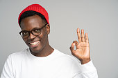 Cheerful playful Afro American man wear red hat in good mood smiling broadly, showing okay gesture over studio grey background. Positive Black male in spectacles advertises a product. Free space