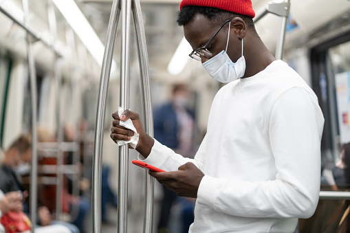 Black millennial man in red hat wearing face mask as protection against covid-19, flu virus, using mobile phone, holding handrail in public transport or subway train through a napkin. New normal.