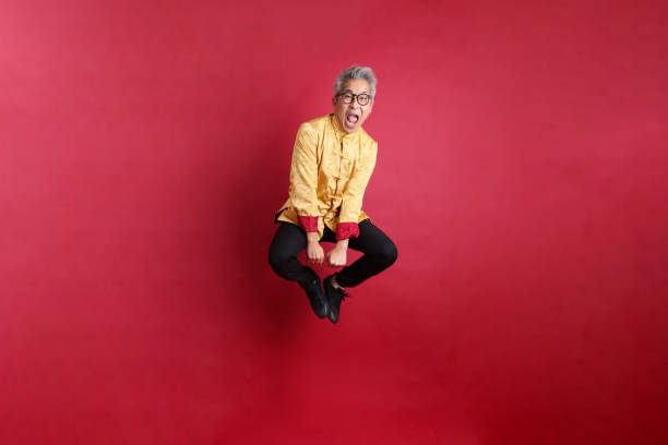 Happy Chinese new year. Asian Chinese energetic senior man wearing golden traditional cheongsam qipao or changshan dress with gesture of jumping isolated on red background.