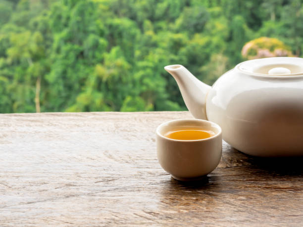 istock Teapots set. White ceramic teapot with hot tea water in small cup preparing on wooden table on natural green mountain view, countryside background with copy space. 1967128923