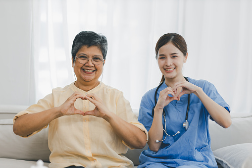 Caregiver nurse Senior patient sit down on sofa and show hands heart symbol. Nurse helping senior Woman