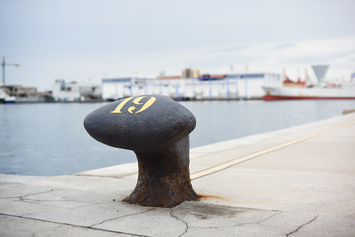 Black marine bollard in harbor with blurred background with ships and industry.