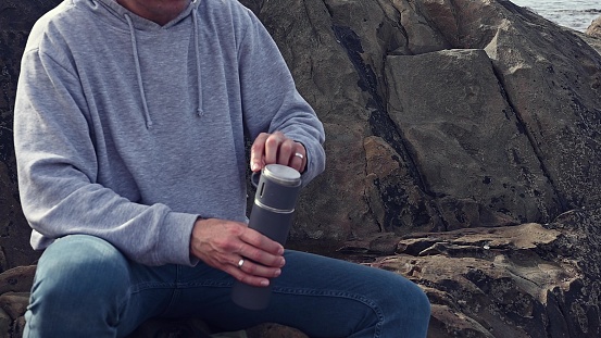 Slow motion young man hiker hipster traveler connecting with nature, opening thermos flask, pouring hot drink into cup, sitting relaxed on a rock by Atlantic ocean. People. Lifestyle. Leisure activity