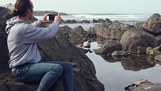 Side view of a Caucasian young adult hipster man photographing sea through smart phone, capturing the beauty of nature, sitting on the rock while beautiful waves splashing and pounding on the headland