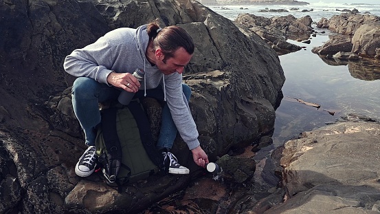 Full size portrait of a Caucasian young adult man tourist, adventurer, hiker, traveler rinsing the stainless steel cup from thermos flask bottle by washing it in sea water, sitting on the rock