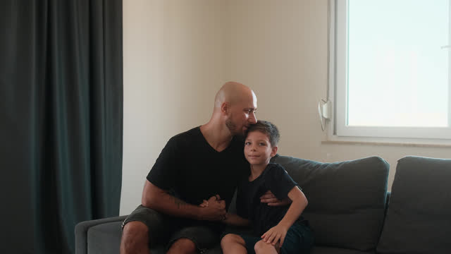 Smiling bald man and young boy sitting on couch, holding hands and interacting with joy.