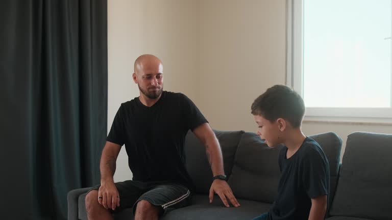 Smiling bald man and young boy sitting on couch, holding hands and interacting with joy.