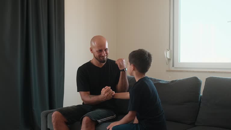 Smiling bald man and young boy sitting on couch, holding hands and interacting with joy.