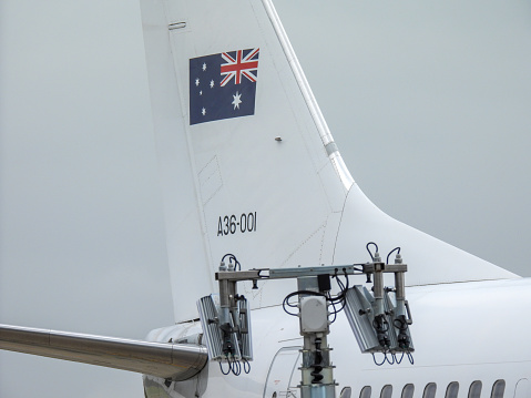 The vertical stabiliser of a Royal Australian Air Force Boeing B737-7DT (BBJ) plane, registration A36-001, used for VIP transport such as the Governor-General and Prime Minister.  She is parked at Sydney Kingsford-Smith Airport waiting to take the Governor-General to Melbourne.  In the foreground is a portable telecommunications tower.  This image was taken from Ross Smith Avenue, Mascot, behind a steel security fence, on a hot, humid and cloudy afternoon on 27 January 2024.