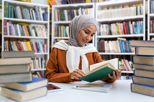 Young beautiful female student studying in the library, female student in hijab smiling among books studying material independently reading literature.