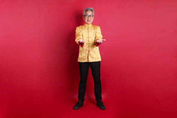 Happy Chinese new year. Asian Chinese energetic senior man wearing golden traditional cheongsam qipao or changshan dress with gesture of pointing isolated on red background.