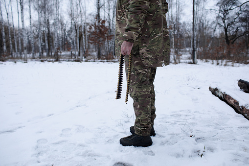 a man in military uniform stands in the snow in the forest and holds a machine gun belt with casings in his hand