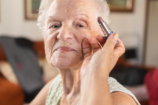 point of view of daughter applying makeup to her mother. applying lipstick to old woman on her lips.happy smiling old woman. femininity and aging process.