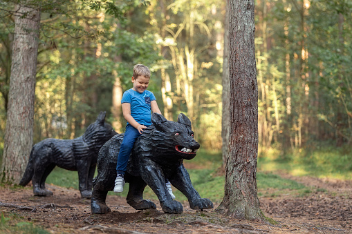 A cheerful smiling boy is riding on a black wolf sculpture on the background of a fairy forest.