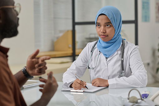 Young female doctor in hijab and uniform attentively listening to her patient and taking notes