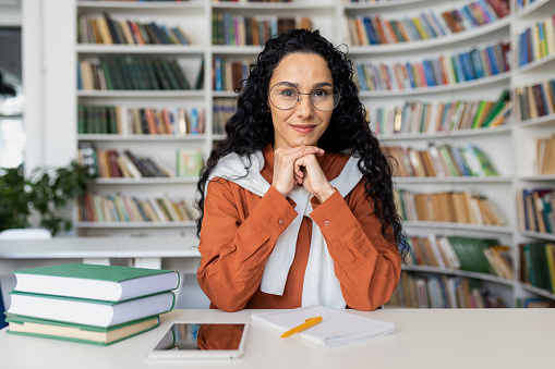 Portrait of a Hispanic female student sitting at a table with textbooks in the classroom, resting her chin on her hands, looking at the camera.