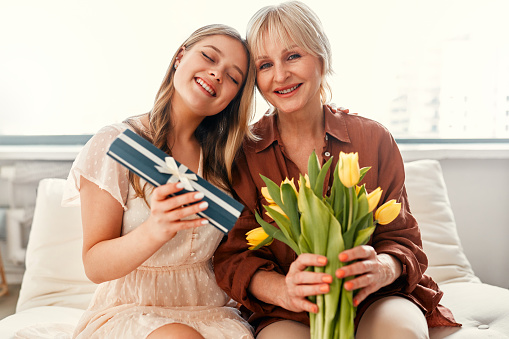 Happy Mother's Day and Women's Day. A daughter congratulating her mother by giving her a bouquet of tulips and a gift while sitting in the living room by the window. Mother's love and care.