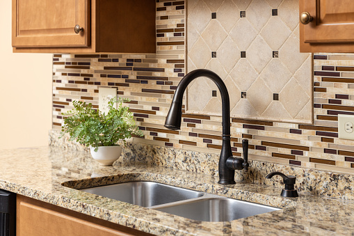 A kitchen faucet detail with wood cabinets, an oil rubbed bronze faucet, quartz countertop, and glass and stone tile backsplash.