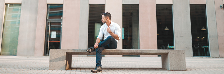 Mature businessman with neat beard wearing white shirt sits on bench on the street of modern city, Panorama. Successful man is resting