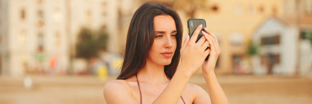 istock Beautiful brown-haired woman with long hair is using mobile phone while standing on the beach, Panorama. Gorgeous girl takes photo on smartphone. 1967543990