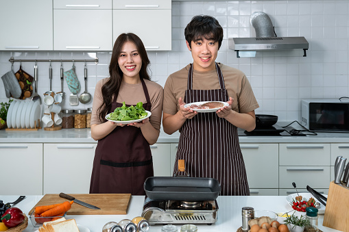 Portrait of young Asian couple male man female woman cheerful standing and preparing food for meal, enjoy cooking, in kitchen at home. Looking at camera.
