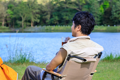 Asian male man sitting on chair, drinking coffee and feeling relax with camping tent beside lake and mountain.