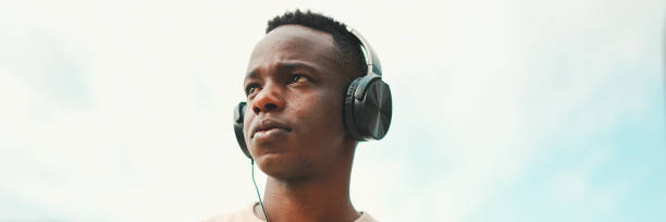 istock Young african student sitting outside of university and taking notes while study online with headphones on blue scy background, Panorama 1967548037