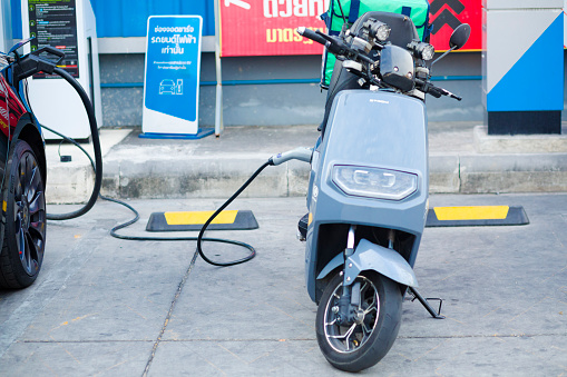 Express delivery EV motor scooter at charger station of PTT gas station which is located at Wanghin Road in Bangkok Ladprao. On scooter is a delivery box of a service company. At side of motorcycle a Tesla car is charged