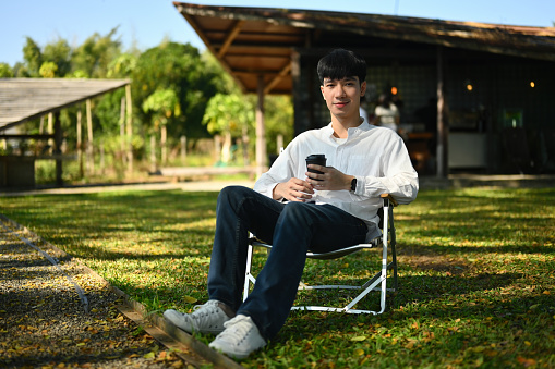 Full-length portrait of a Young Asian man sitting on a lawn chair and holding a cup of coffee, outdoors in the morning with a sunkissed face.