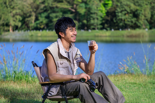 Young Asian male man sitting on chair, drinking coffee, feeling relax and raising hands, during camping beside lake and mountain.