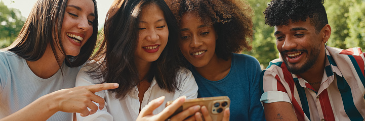 Happy, smiling multiethnic young people at picnic on summer day outdoors, Panorama. Group of friends talking, using cellphone while relaxing in the park at picnic