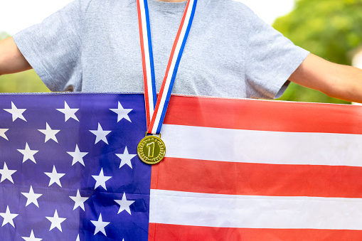 Triumphant American male athlete proudly displays a gold medal, symbolizing victory. Holding the American flag, he embodies national pride during sporting events.