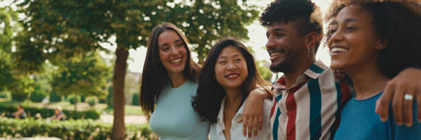 istock Happy multiethnic young people walk embracing on summer day outdoors, Panorama. Group of friends are talking and laughing merrily while walking along path in city park 1967565440