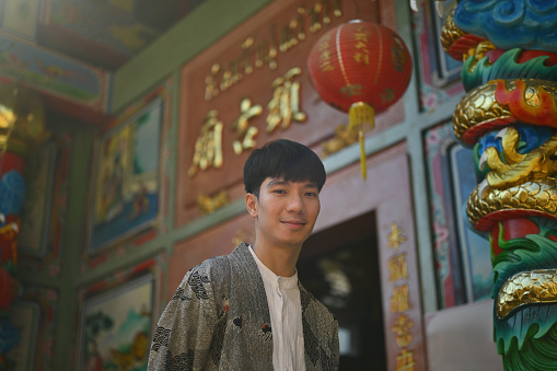 Portrait of a happy and smiling young Asian man wearing a robe standing in front of the entrance to a Chinese shrine.