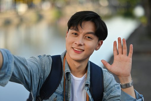 Handsome young Asian traveler smiling and waving at mobile camera, Travel Influencer and Solo traveler concept.