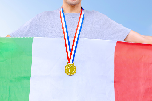 Triumphant Italian male athlete proudly displays a gold medal, symbolizing victory. Holding the flag of Italy, he embodies national pride during sporting events.