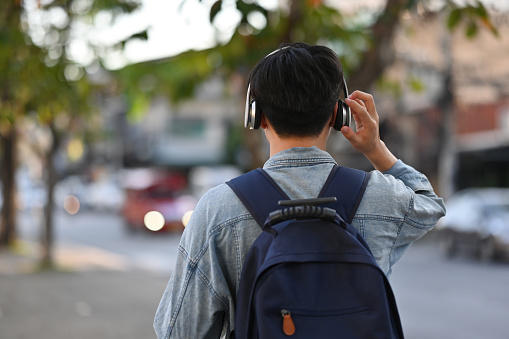 Rear view of Asian backpacker male walking on footpath while listening a music from headphones in the urban city, Solo travel concept.