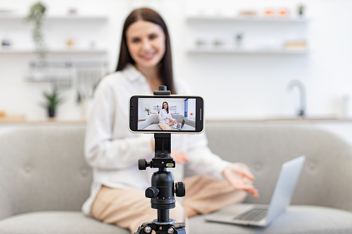 Young caucasian woman in with laptop sitting on comfortable sofa in kitchen. Front view of cheerful content maker in casual wear recording video to followers on modern smartphone.