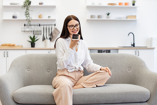 Beautiful woman relaxing on couch and using modern smartphone for recording conversation. Caucasian brunette using modern devices enjoying break from work for exchanging of audio messages on weekend.