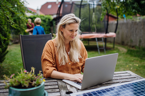 Businesswoman, freelancer or manager working outdoors in the garden, garden homeoffice. Remote work from backyard. Laptop is charging with solar charger. Concept of working remotely.